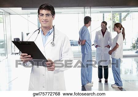 Stock Photo - Doctor holding a clipboard with his colleagues discussing in the background. Fotosearch