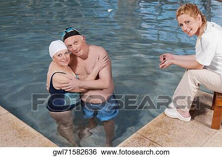 Stock Photograph - Portrait of a couple hugging in a swimming pool with a health worker at poolside. Fotosearch
