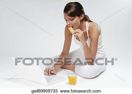Stock Image - Woman eating a snack and working on a laptop. Fotosearch