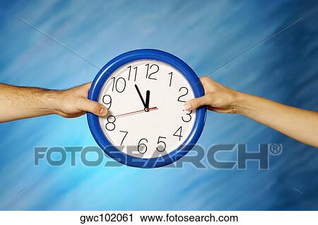 Stock Image - Close-up of a man and a woman's hands holding a wall clock. Fotosearch