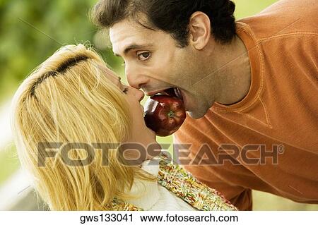 Stock Image - Close-up of a young woman and a mid adult man biting an apple together. Fotosearch