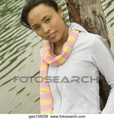 Stock Photo - Portrait of a young woman leaning against a tree trunk by the lake. Fotosearch
