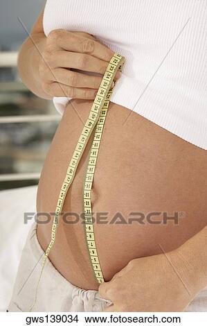 Picture - Mid section view of a pregnant woman measuring her abdomen with a measuring tape. Fotosearch