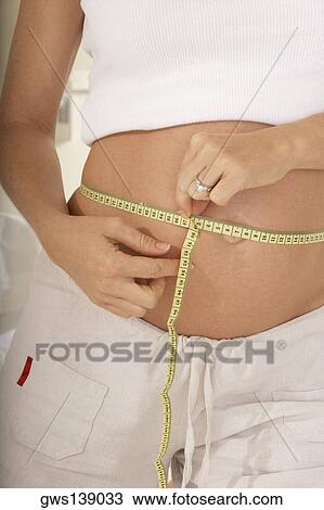 Stock Image - Mid section view of a pregnant woman measuring her abdomen with a measuring tape. Fotosearch