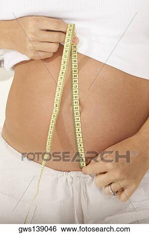 Stock Photograph - Mid section view of a pregnant woman measuring her abdomen with a measuring tape. Fotosearch