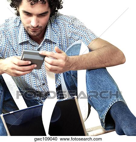 Stock Image - Young man sitting in front of a laptop holding a calculator. Fotosearch