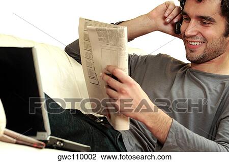 Stock Image - Close-up of a young man reading a newspaper and talking on a mobile phone. Fotosearch