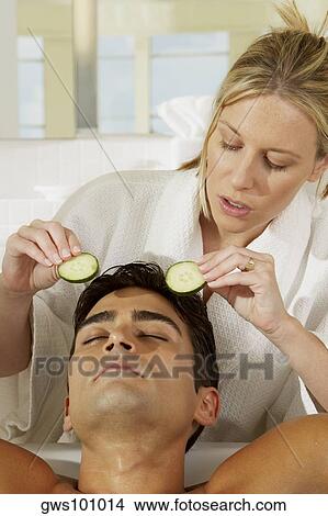 Picture - Close-up of a young woman putting cucumber slices on a young man's eye. Fotosearch