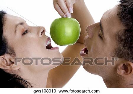 Stock Image - Close-up of a young woman holding a green apple above her mouth and a young man's mouth. Fotosearch