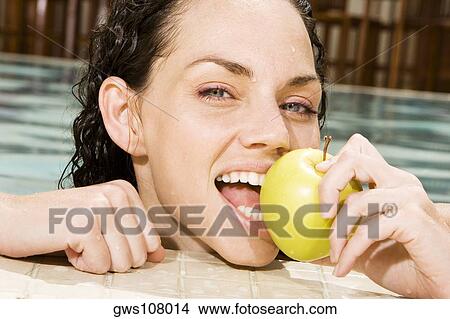 Picture - Portrait of a young woman eating a green apple in a swimming pool. Fotosearch