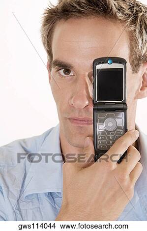 Portrait of a young man holding a mobile phone in front of his eye View Large Photo Image Picture - Portrait of a young man holding a mobile phone in front of his eye. Fotosearch