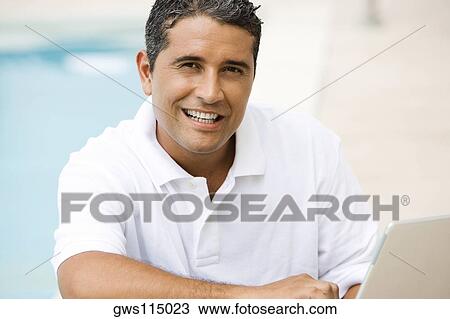 Stock Image - Portrait of a mid adult man using a laptop at the poolside. Fotosearch