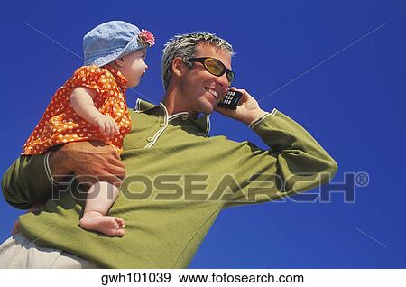 Stock Photo - Low angle view of a mature man carrying a baby girl and talking on a mobile phone. Fotosearch