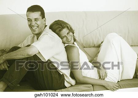 Picture - Side profile of a young couple sitting back to back on a couch and smiling. Fotosearch