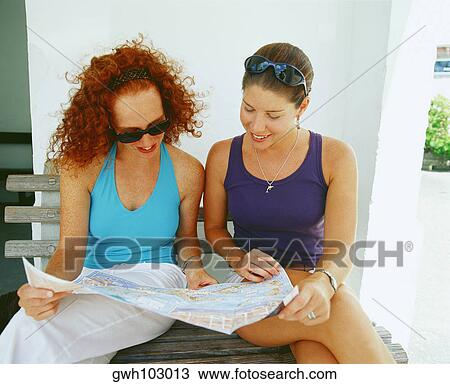 Stock Image - Close-up of two young women sitting on a bench and reading a newspaper, Bermuda. Fotosearch