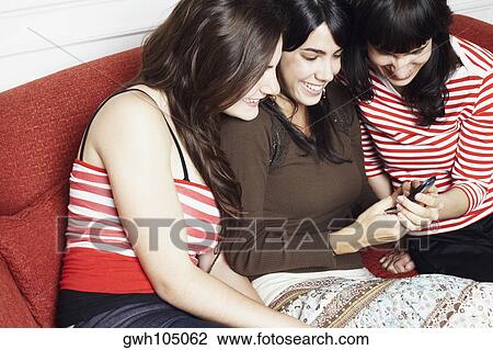 Two young women and a mid adult woman sitting on a couch and looking at a mobile phone View Large Photo Image Stock Image - Two young women and a mid adult woman sitting on a couch and looking at a mobile phone. Fotosearch