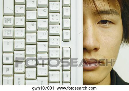 Stock Image - Portrait of a young man covering his face with a computer keyboard. Fotosearch