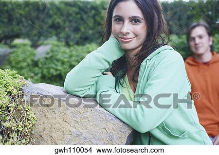 Portrait of a young woman leaning against a stone with a mid adult man in the background View Large Photo Image Picture - Portrait of a young woman leaning against a stone with a mid adult man in the background. Fotosearch