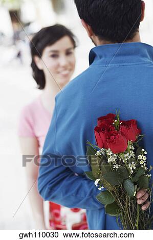 Stock Image - Young man hiding a bunch of roses behind his back with a young woman in front of him. Fotosearch