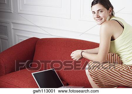 Portrait of a young woman sitting on a couch's arm in front of a laptop View Large Photo Image Stock Image - Portrait of a young woman sitting on a couch's arm in front of a laptop. Fotosearch