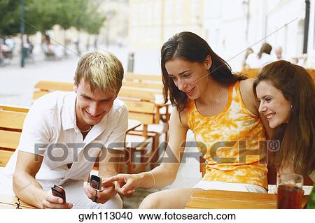 Young man holding two mobile phones with two young women sitting beside him View Large Photo Image Stock Image - Young man holding two mobile phones with two young women sitting beside him . Fotosearch