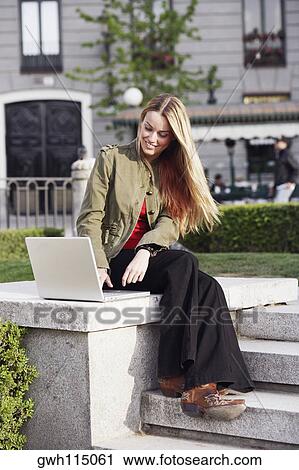 Young woman sitting on a ledge and using a laptop View Large Photo Image Stock Image - Young woman sitting on a ledge and using a laptop . Fotosearch