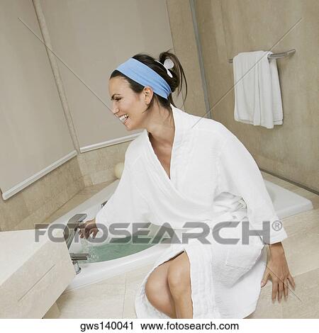 Stock Image - Young woman sitting near a bathtub checking the water temperature. Fotosearch
