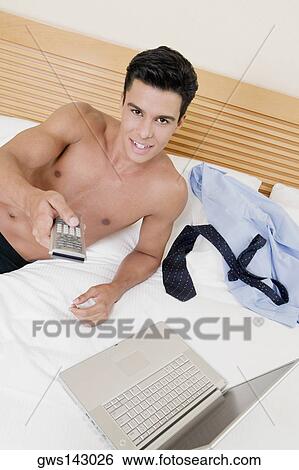 Portrait of a young man lying on the bed using a remote control with a laptop in front of him View Large Photo Image Stock Photograph - Portrait of a young man lying on the bed using a remote control with a laptop in front of him. Fotosearch