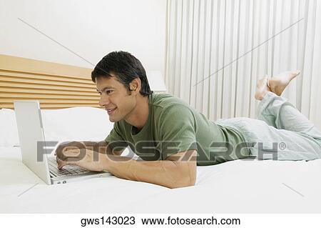 Stock Image - Side profile of a mid adult man lying on the bed and using a laptop. Fotosearch