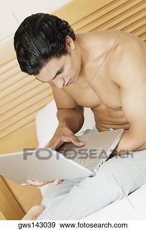 Side profile of a young man sitting on the bed and using a laptop View Large Photo Image Stock Photo - Side profile of a young man sitting on the bed and using a laptop. Fotosearch