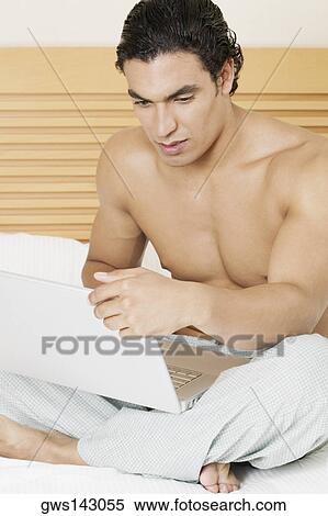 Young man sitting on the bed and using a laptop View Large Photo Image Stock Photography - Young man sitting on the bed and using a laptop. Fotosearch
