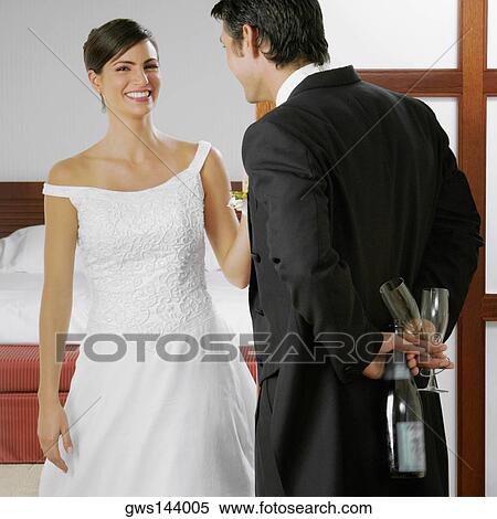 Stock Photography - Groom looking at his bride and holding a bottle and glasses behind his back. Fotosearch