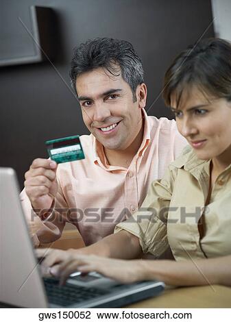 Stock Image - Portrait of a mature man holding a credit card and a mid adult woman using a laptop near him. Fotosearch