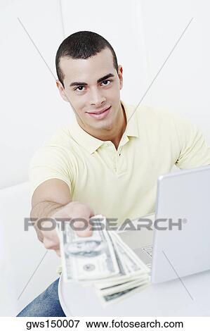 Portrait of a young man holding dollar bills in front of a laptop View Large Photo Image Stock Photo - Portrait of a young man holding dollar bills in front of a laptop. Fotosearch