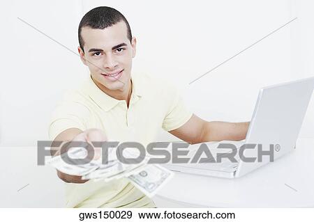 Portrait of a young man holding dollar bills in front of a laptop View Large Photo Image Stock Photo - Portrait of a young man holding dollar bills in front of a laptop. Fotosearch
