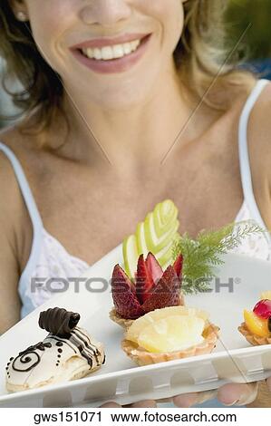 Stock Image - Close-up of a young woman holding assorted tarts with an eclair in a platter. Fotosearch