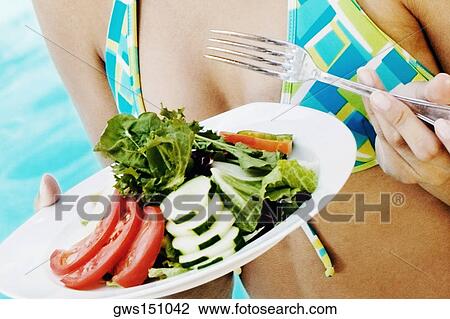 Stock Image - Mid section view of a woman holding a plate of vegetable salad and fork. Fotosearch