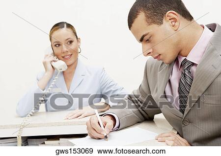 Stock Photo - Close-up of a businessman writing on paper and a female receptionist talking on the telephone beside him. Fotosearch