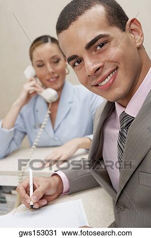 Stock Image - Portrait of a businessman smiling with a female receptionist talking on the telephone behind him. Fotosearch