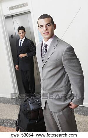 Portrait of a businessman with luggage and a businessman standing in front of an elevator View Large Photo Image Picture - Portrait of a businessman with luggage and a businessman standing in front of an elevator. Fotosearch