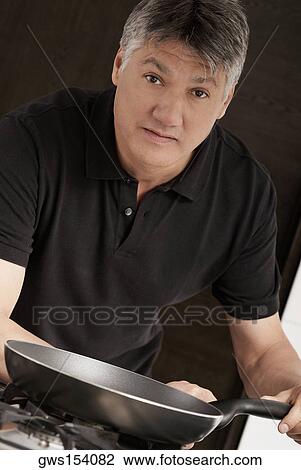 Stock Image - Portrait of a senior man holding a frying pan in the kitchen. Fotosearch