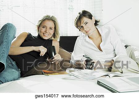 Stock Photo - Portrait of two mid adult women holding cups of coffee on the bed. Fotosearch