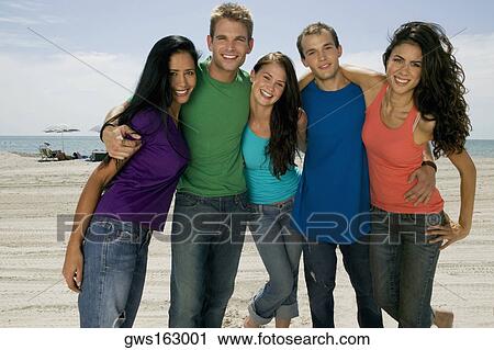 Stock Image - Portrait of three young women and two young men standing on the beach. Fotosearch
