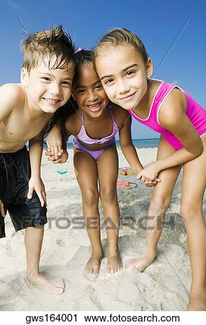 Stock Image - Portrait of two girls and a boy standing on the beach and smiling. Fotosearch