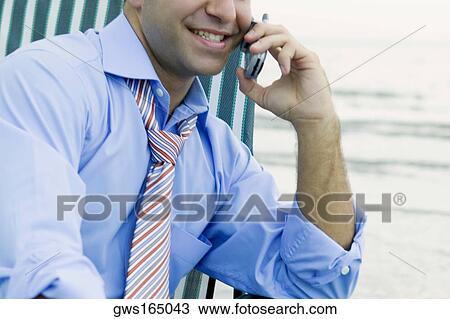 Stock Image - Close-up of a young man sitting on a chair on the beach and talking on a mobile phone. Fotosearch