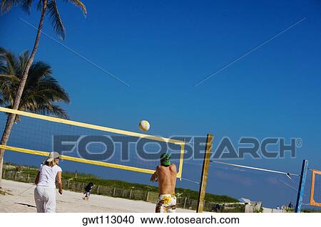 Stock Image - Rear view of a man with a woman playing beach volleyball. Fotosearch