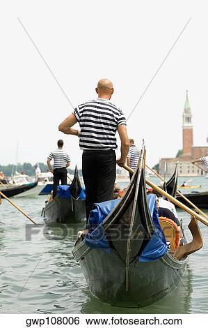 Rear view of three gondoliers standing on gondolas, Venice, Veneto, Italy View Large Photo Image Stock Photograph - Rear view of three gondoliers standing on gondolas, Venice, Veneto, Italy. Fotosearch