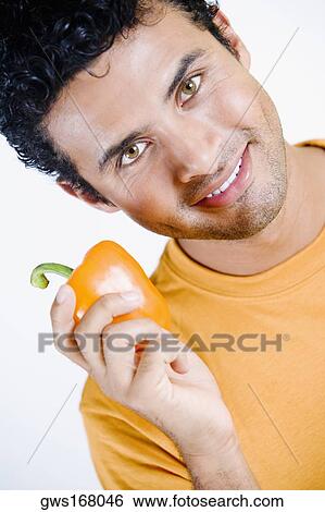 Stock Photograph - Portrait of a young man holding an orange bell pepper and smiling. Fotosearch