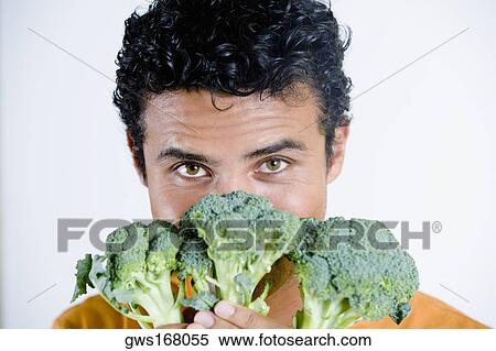 Stock Photography - Portrait of a young man holding broccoli in front of his face. Fotosearch
