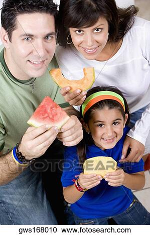 Stock Image - Portrait of a young woman and a mid adult man with their daughter holding slices of fruits. Fotosearch